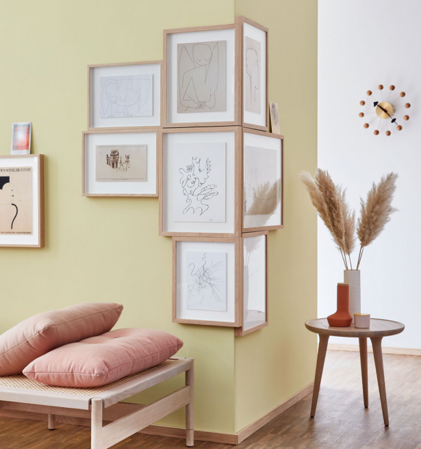Contemporary living room corner decorated with a cluster of framed line drawings on a pale yellow wall, complemented by a wooden bench with pink cushions, a small round side table, and a vase with pampas grass, creating a modern and simplistic interior de